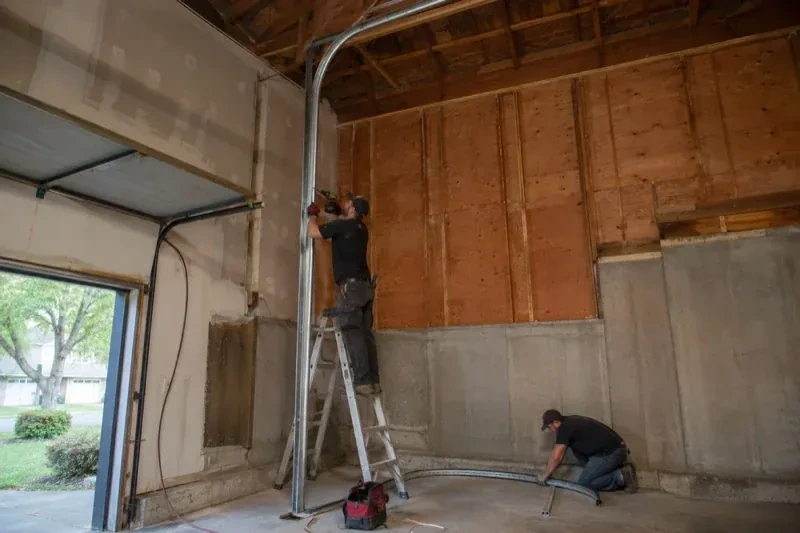 One person on a stepladder working beside a tall curved garage-door track while another kneels beside loose track sections in an unfinished garage. — High-Lift Garage Door Conversion, Uxbridge